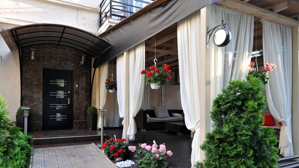 A hotel entrance with a dark marble facade, a black door with a metal handle, and a small outdoor seating area with white curtains and hanging flower baskets. - Reno (Odesa)