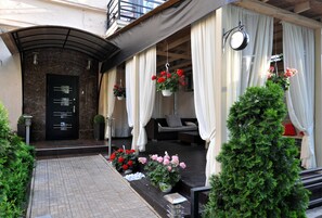 A hotel entrance with a dark marble facade, a black door with a metal handle, and a small outdoor seating area with white curtains and hanging flower baskets. - Reno (Odesa)