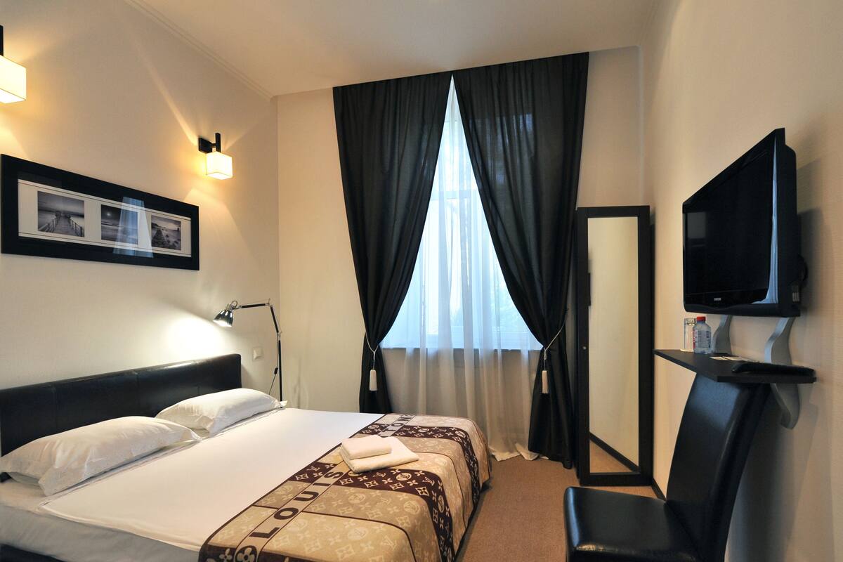 A hotel room with a bed, a desk with a chair, a television, and a window with curtains.