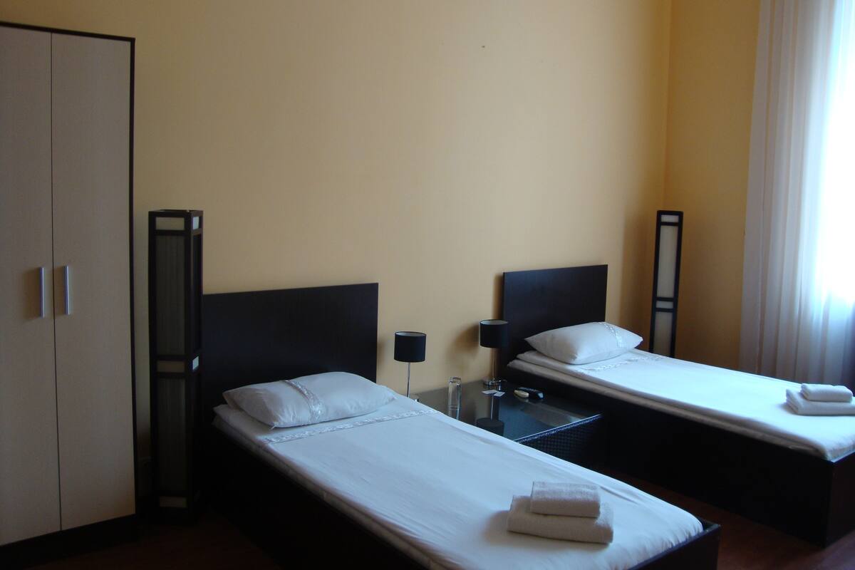 A hotel room with two single beds, a wardrobe, a bedside table with a lamp, and a window with curtains.