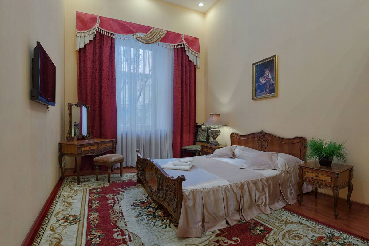 A bedroom with a wooden bed, a desk, a chair, a mirror, a nightstand with a lamp, and a potted plant.