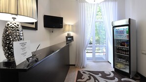 A hotel reception area with a counter, a television, a lamp, a refrigerator stocked with beverages, and a view of greenery outside. - Reno (Odesa)