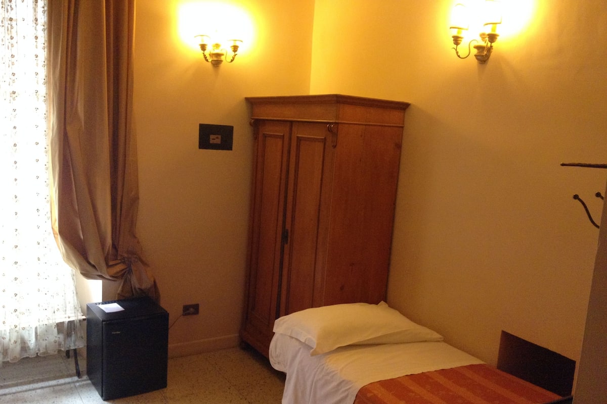 A small, dimly lit room with a single bed, a wooden wardrobe, and a desk with a computer.