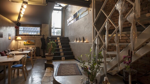 Un comedor con mesas y sillas de madera, una escalera con barandilla de metal y vistas al exterior a través de una ventana.