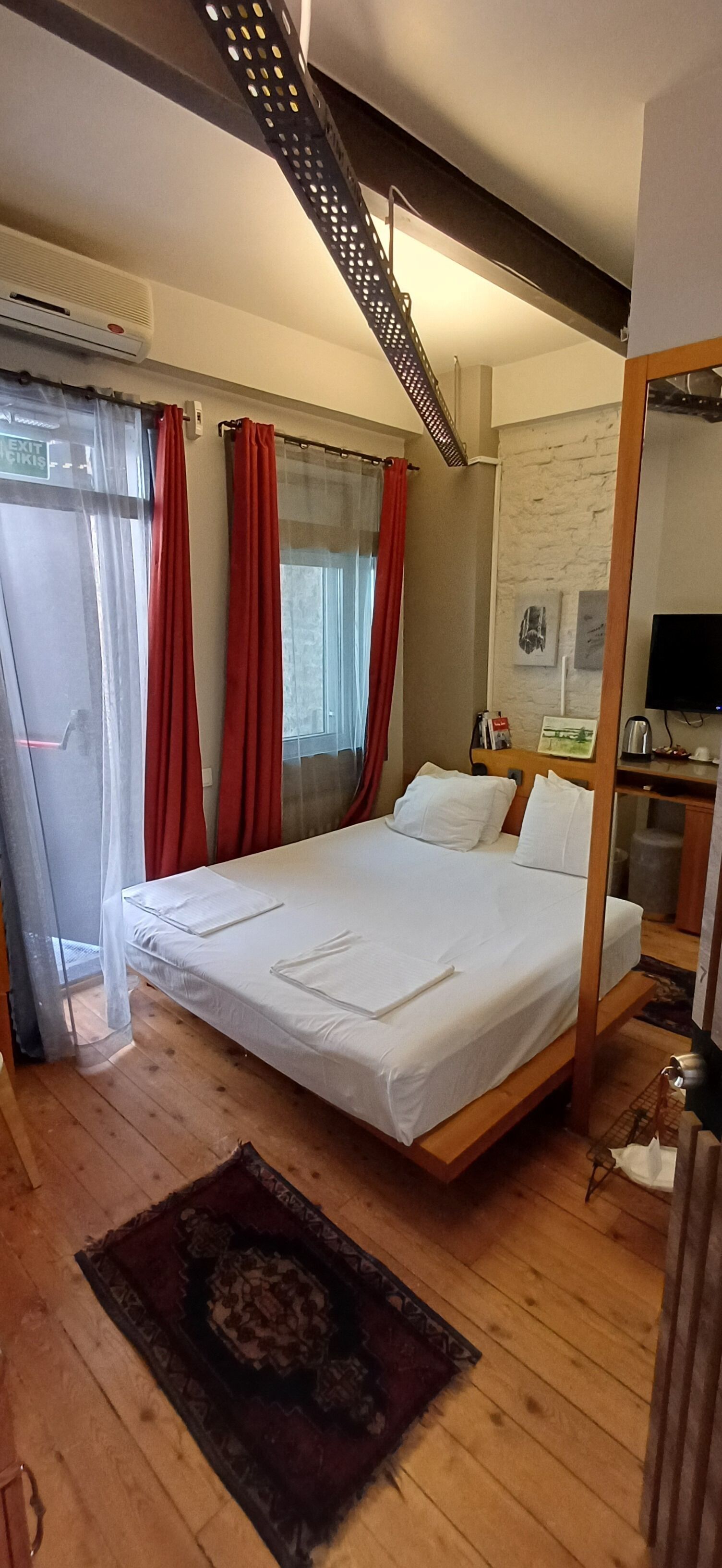 Une chambre à coucher avec un lit, des rideaux, un plancher en bois, un miroir et un téléviseur.