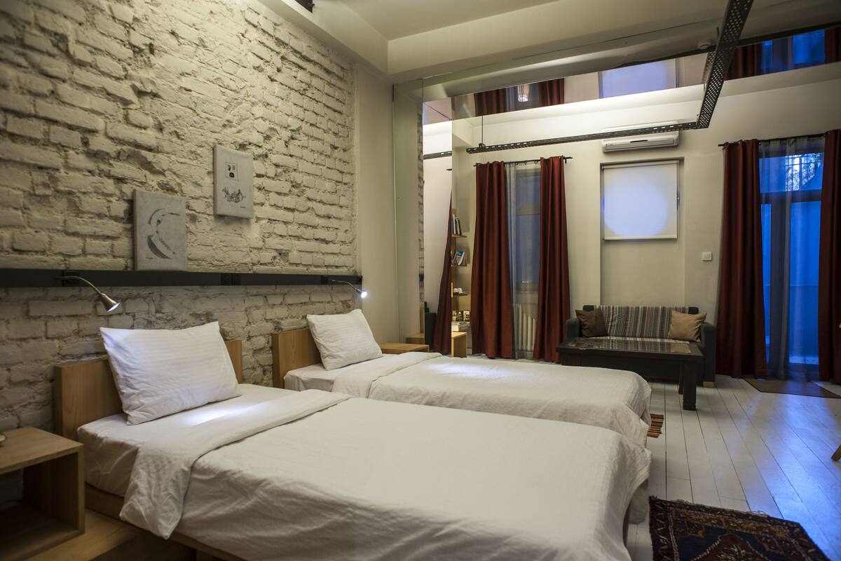 A room with two beds, a brick wall, a sofa, and a large window with curtains.