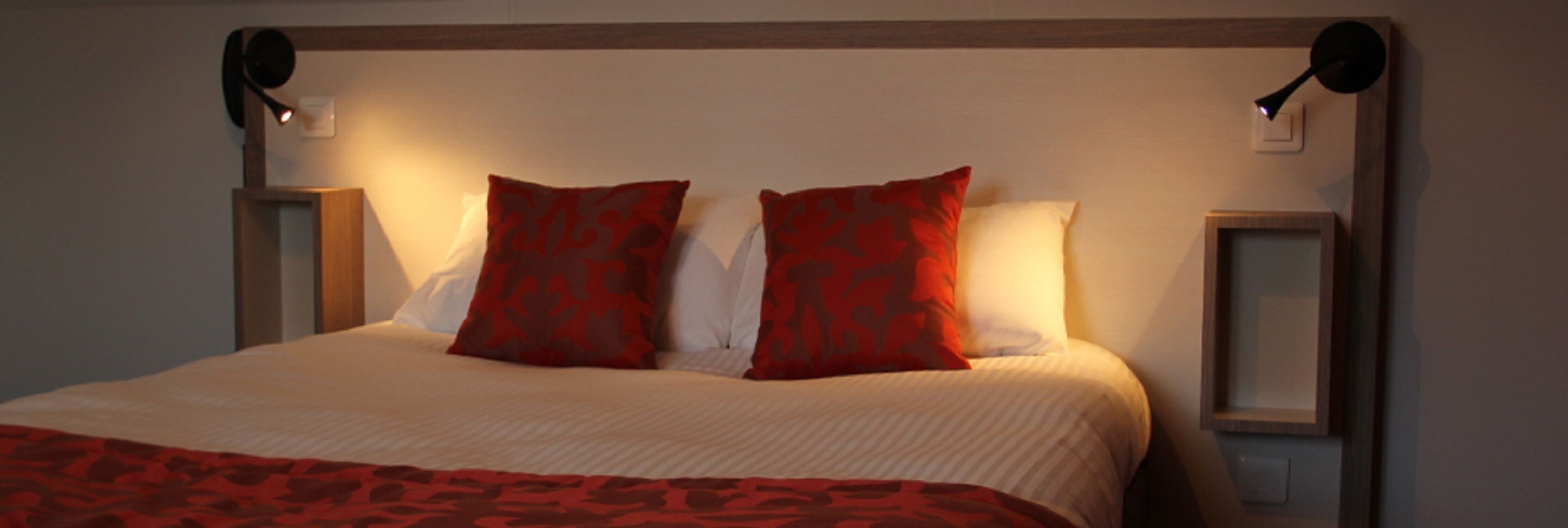A neatly made bed with a red and white patterned bedspread, two red pillows, and two bedside lamps on either side.