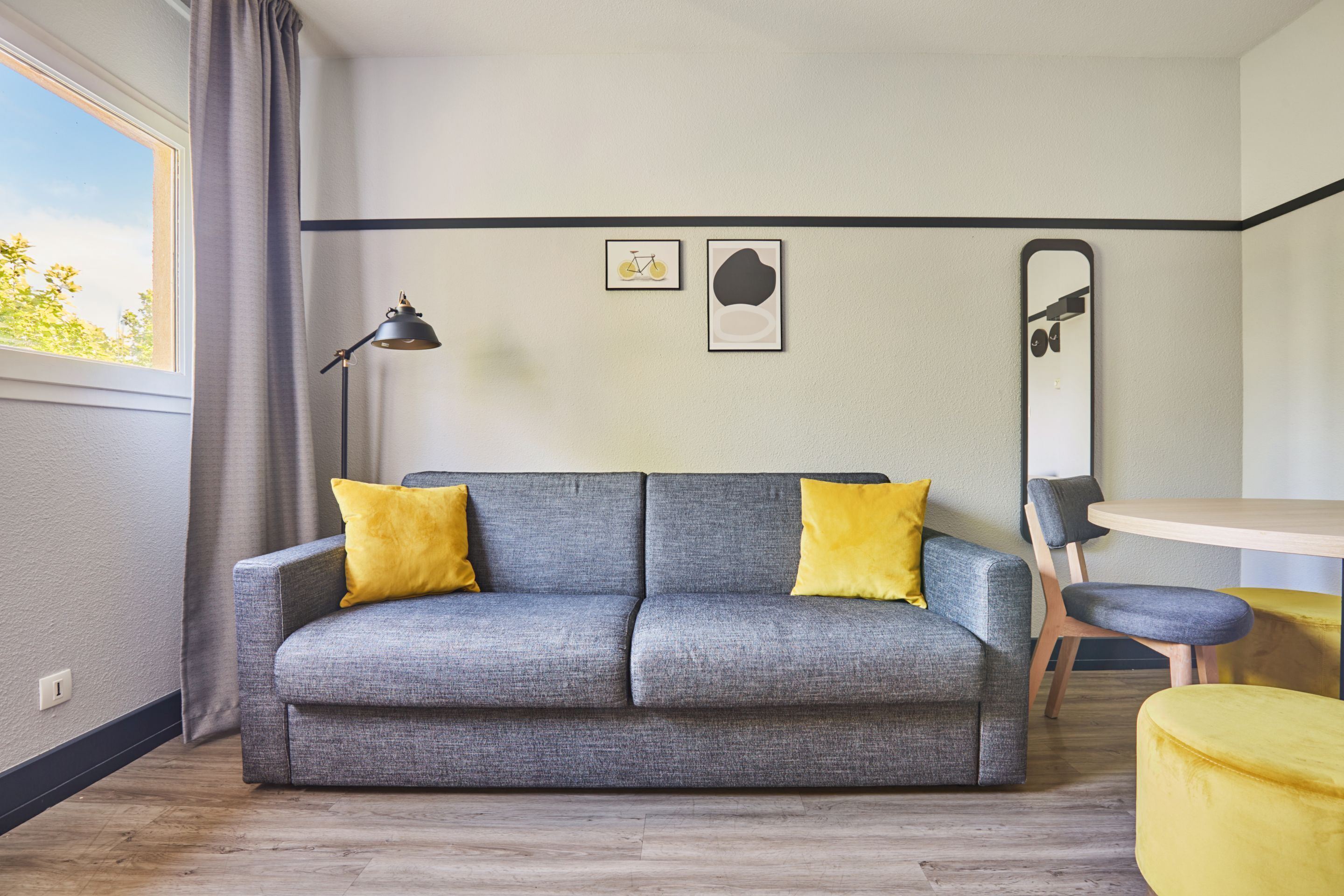 A modern living room with a grey sofa, two yellow cushions, a black floor lamp, a round table, and a mirror on the wall.