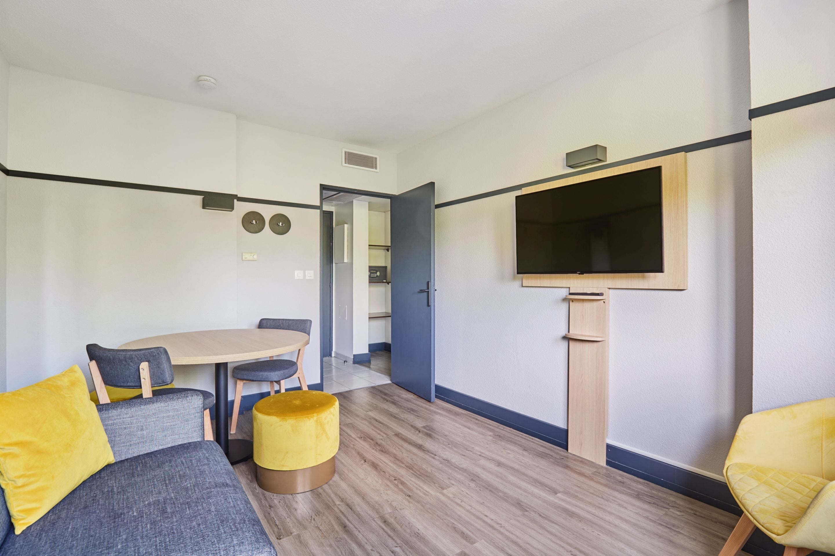 Une chambre d’hôtel moderne, équipée d’un canapé, d’une table ronde, d’un pouf jaune et d’un téléviseur à écran plat fixé au mur.