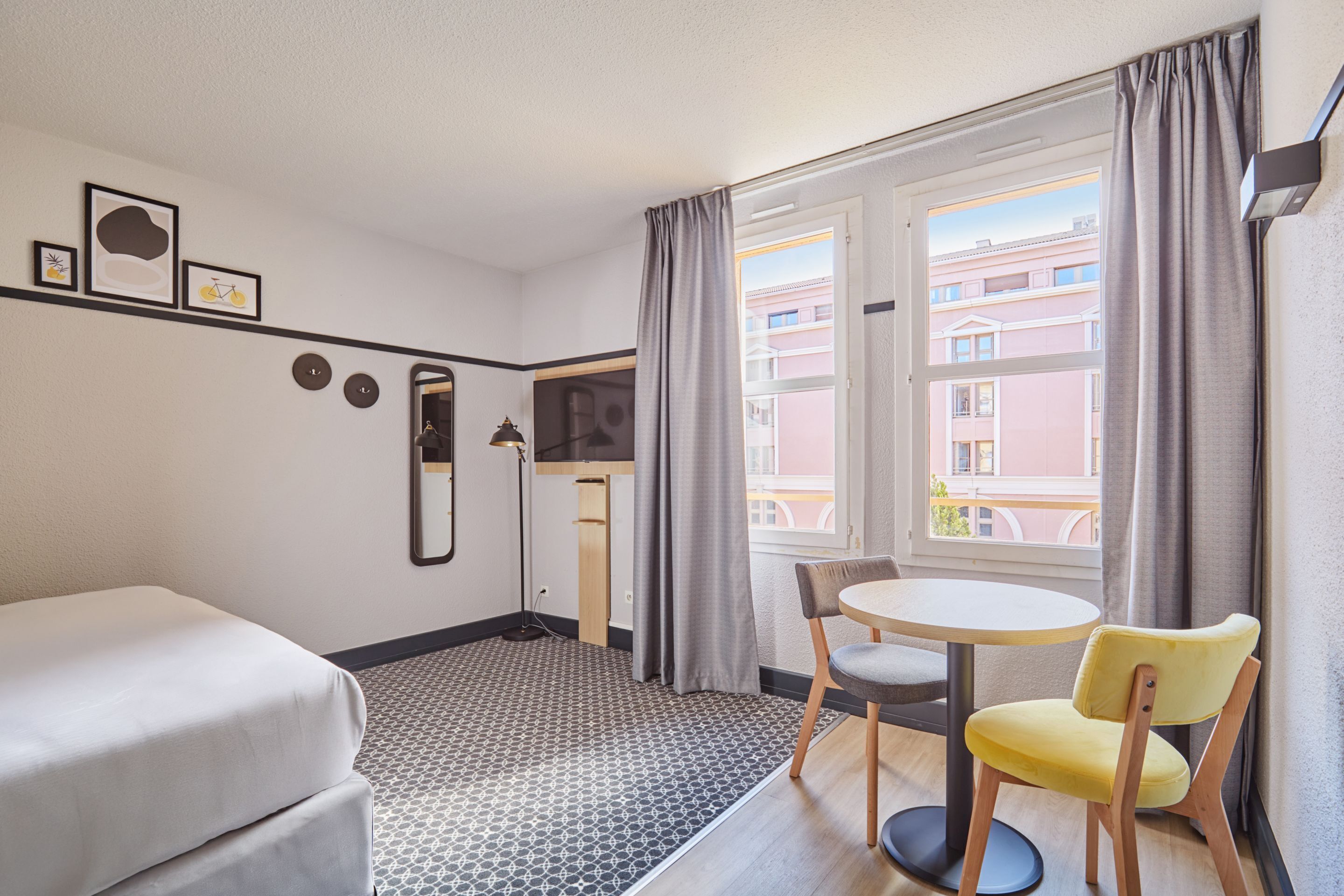 Une chambre d’hôtel avec un lit, une petite table et deux chaises, un miroir et une fenêtre avec des rideaux.