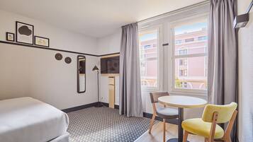 Une chambre d’hôtel avec un lit, une petite table et deux chaises, un miroir et une fenêtre avec des rideaux.