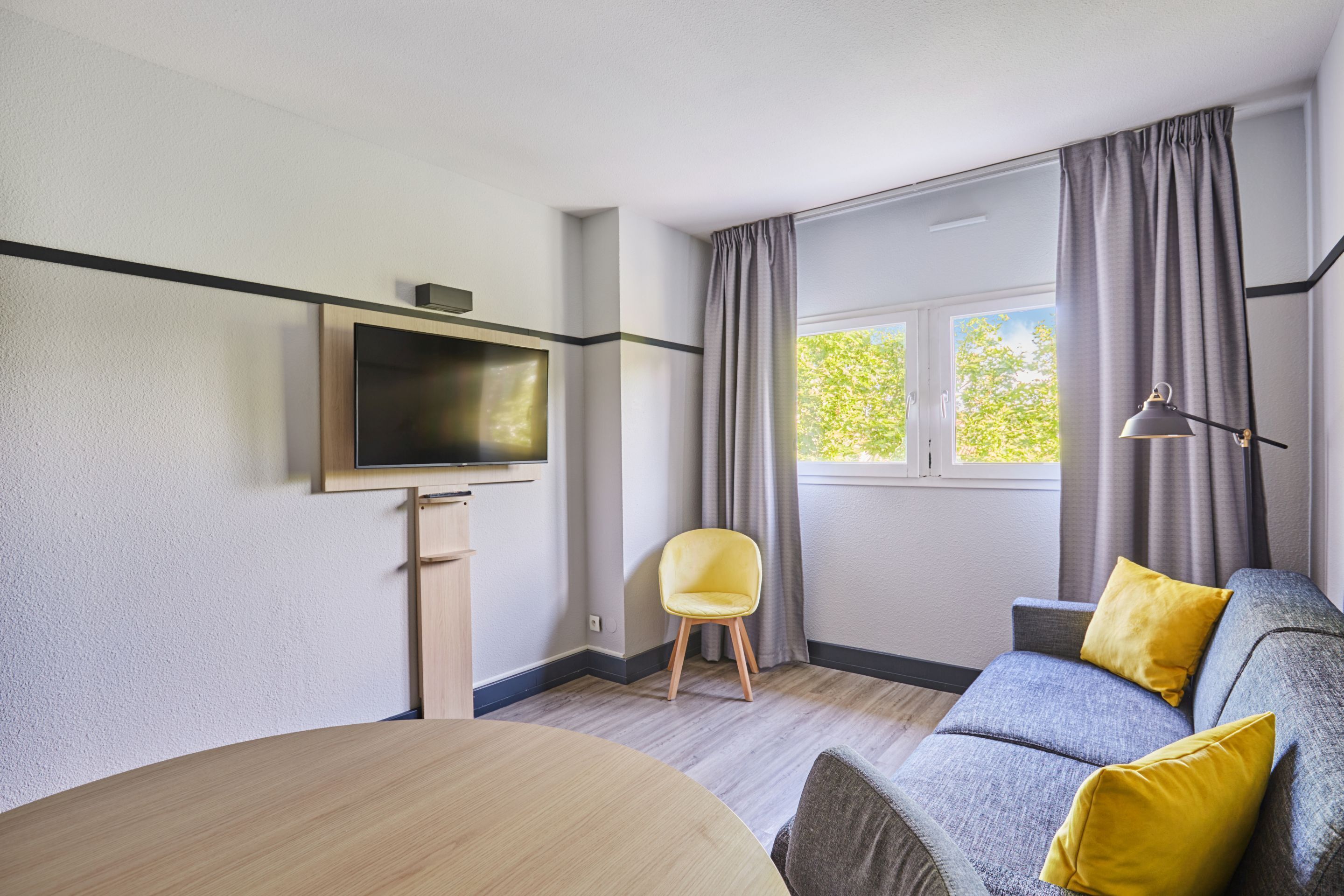 A modern living room with a flat-screen TV, a round dining table, a sofa with yellow cushions, and a window with curtains.