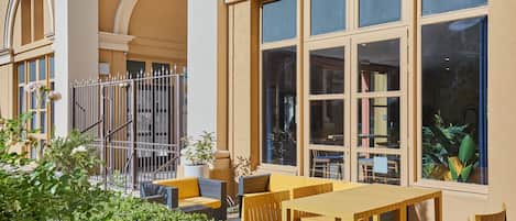 An outdoor seating area with tables and chairs, surrounded by greenery and a building facade with large windows.