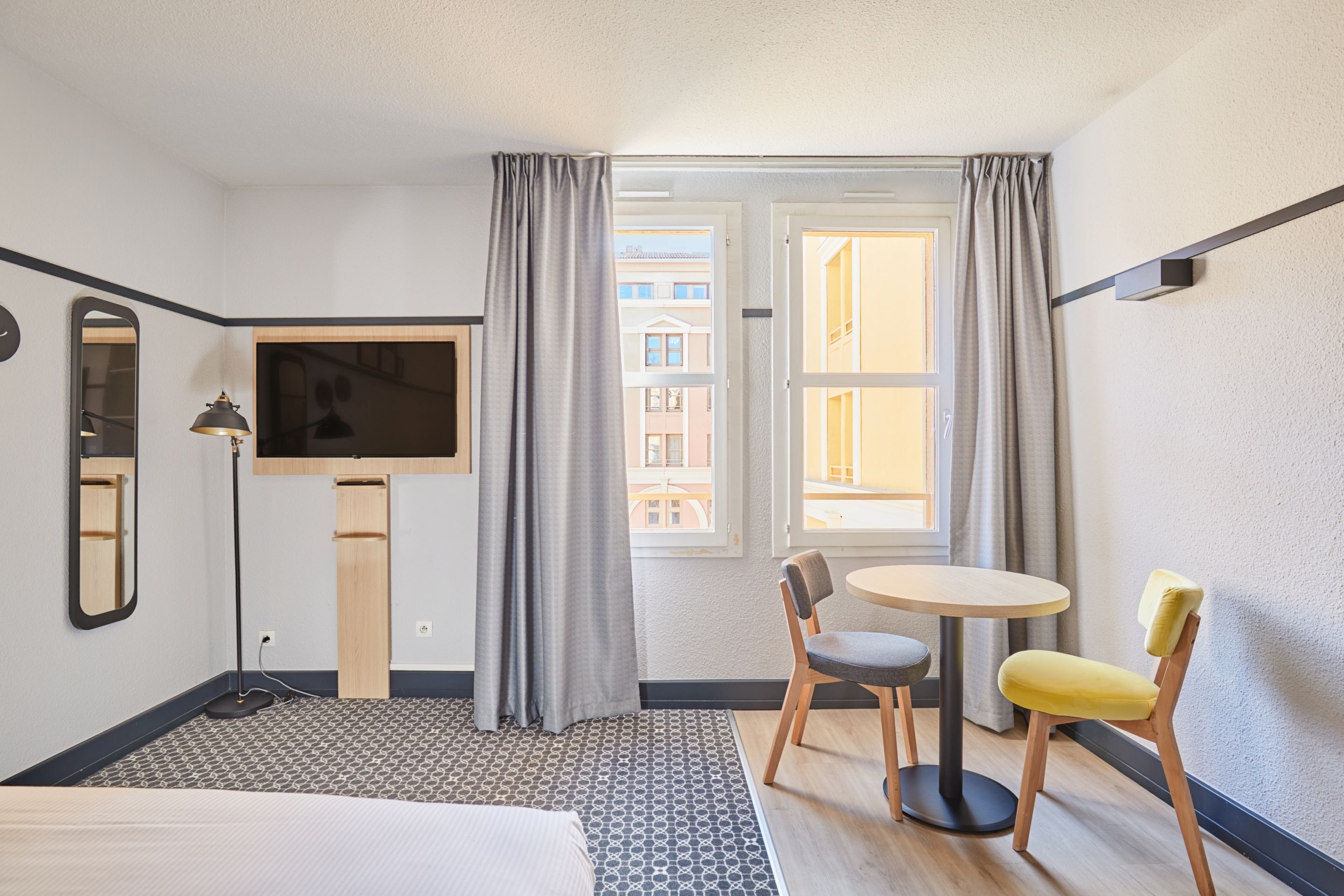 Quarto de hotel com uma cama, televisão, uma mesinha com duas cadeiras, janela com cortinas e um espelho.