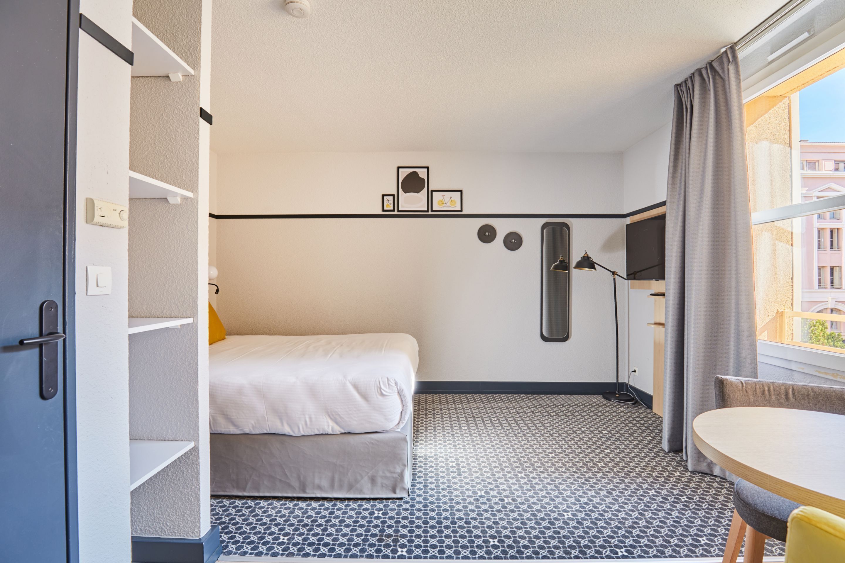 A compact hotel room with a bed, a desk, a chair, a TV, and a window with curtains.