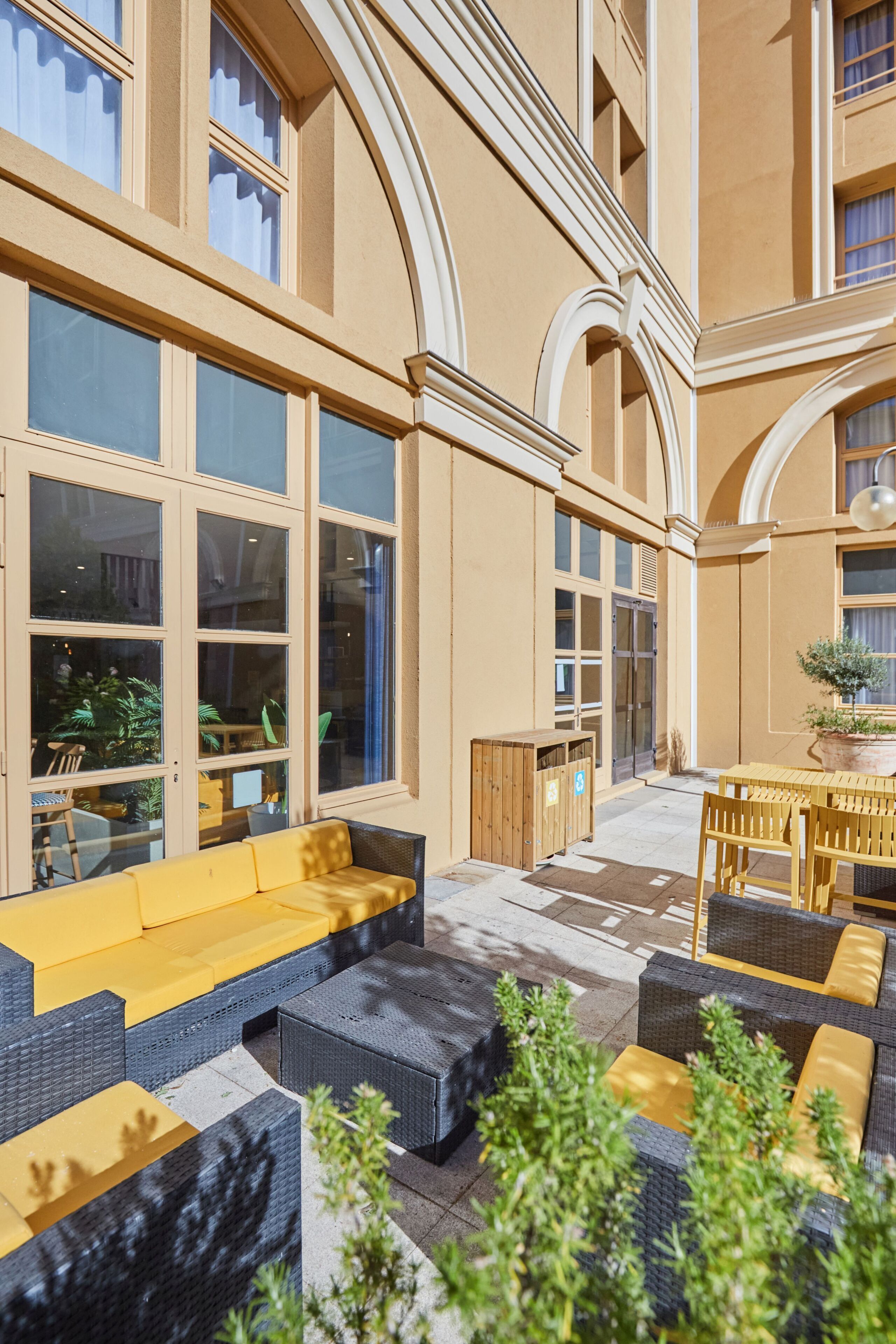 A spacious outdoor seating area with multiple sofas and tables, surrounded by large windows and potted plants.
