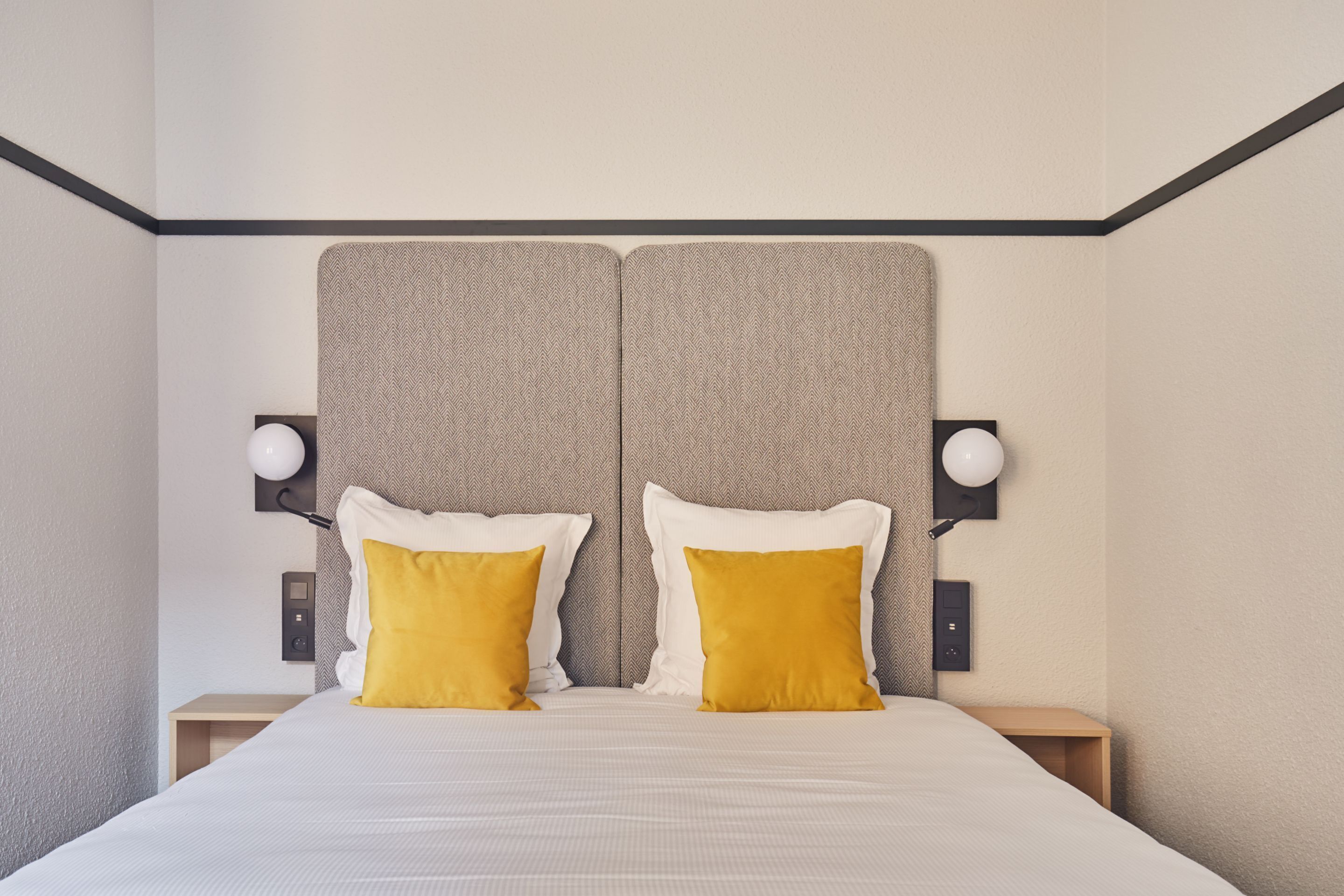 Un lit double avec deux oreillers blancs et deux oreillers de couleur jaune moutarde, flanqué de deux tables de chevet avec des lampes, dans une chambre dont la tête de lit est texturée et dont les murs sont de couleur unie.