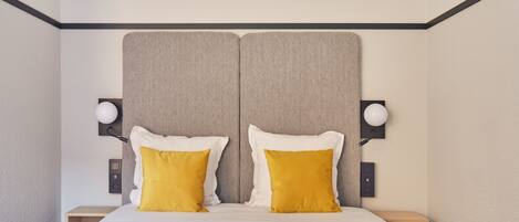 Un lit double avec deux oreillers blancs et deux oreillers de couleur jaune moutarde, flanqué de deux tables de chevet avec des lampes, dans une chambre dont la tête de lit est texturée et dont les murs sont de couleur unie.