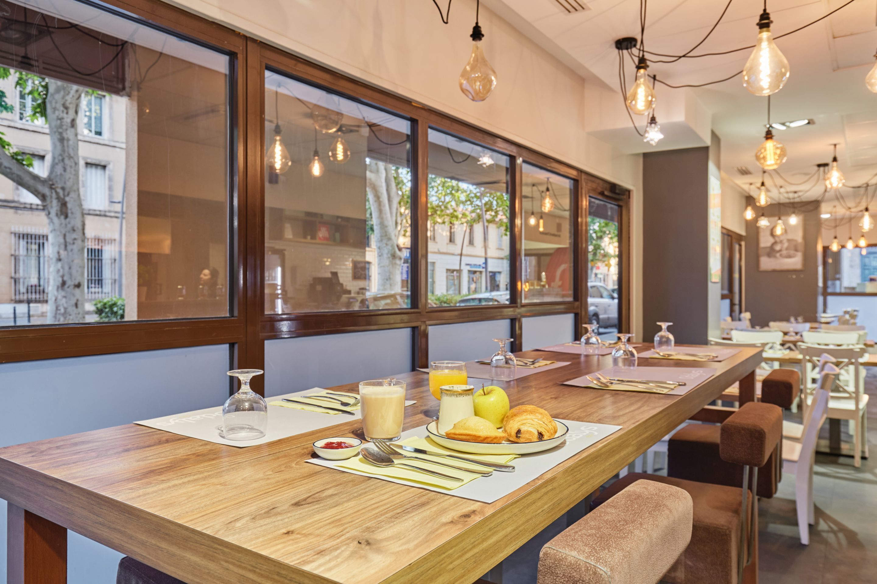 A breakfast setup with a wooden table, breakfast items, and a view of the street through large windows.