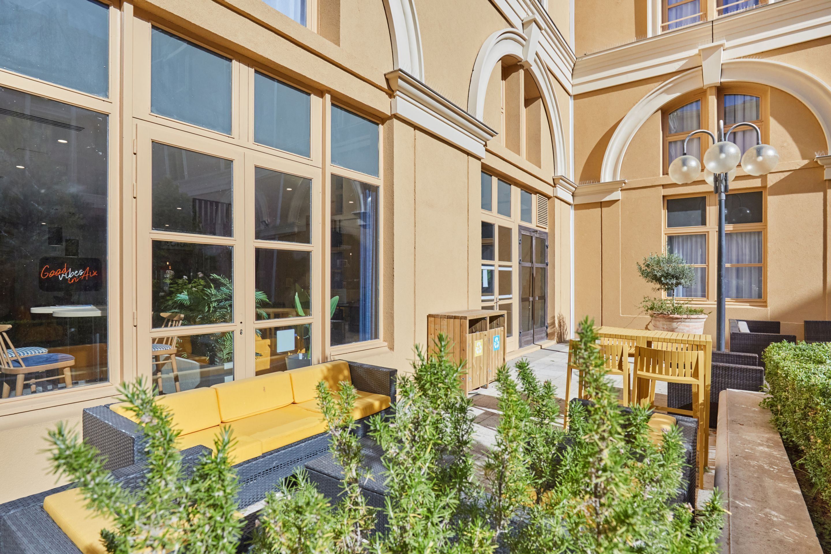 An outdoor seating area with a yellow sofa and wooden tables, surrounded by greenery and a building with large windows and arched doorways.
