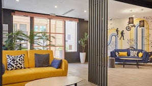 A modern lobby with a yellow sofa, a round table, and a blue sofa with yellow cushions. There are potted plants and a mural with people hiking. - Odalys City Apartment Hotel - Aix en Provence Centre Palais des Congrès (Aix-en-Provence)