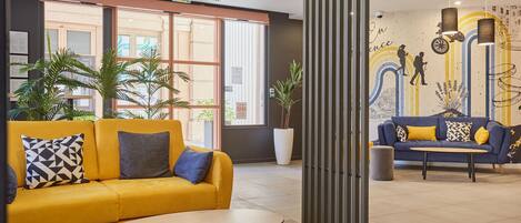 A modern lobby with a yellow sofa, a round table, and a blue sofa with yellow cushions. There are potted plants and a mural with people hiking.