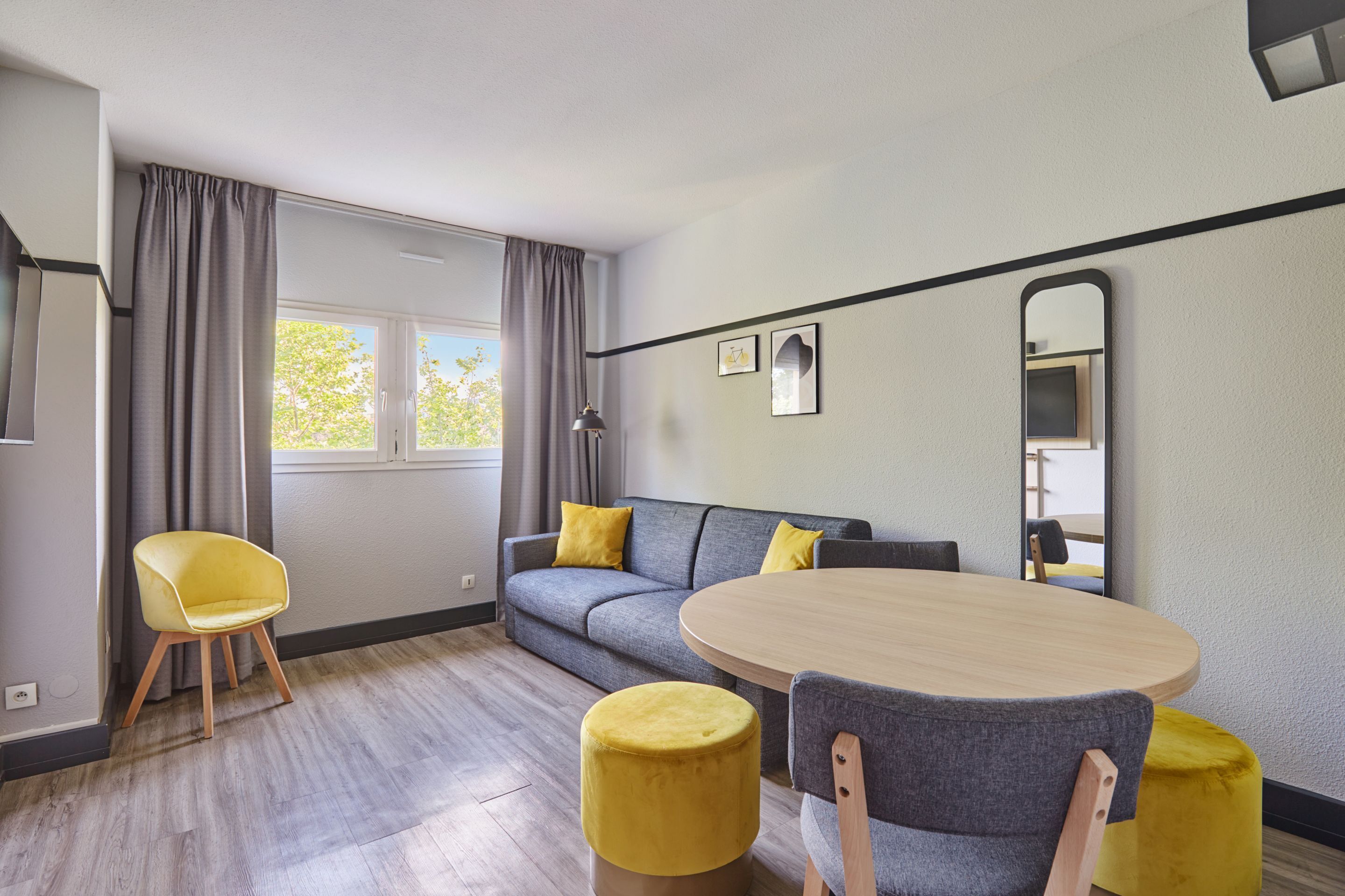 Ein modernes Wohnzimmer mit grauem Sofa, gelben Kissen, einem runden Holztisch und passenden Hocker.