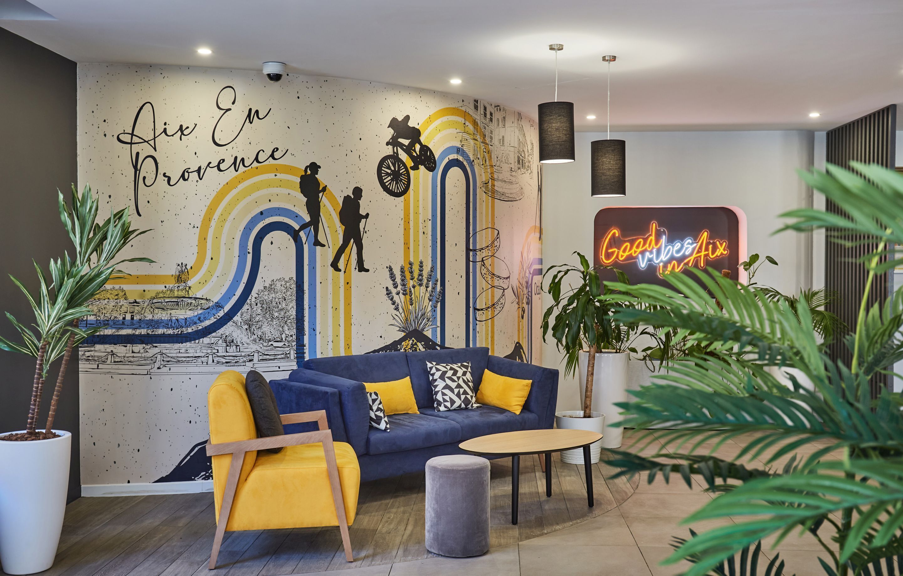 Een moderne woonkamer met een blauwe bank, een gele fauteuil en een ronde salontafel. Een muurschildering met 'Aix En Provence' en een neonbord met de tekst 'Good vibes Aix' sieren de wand.