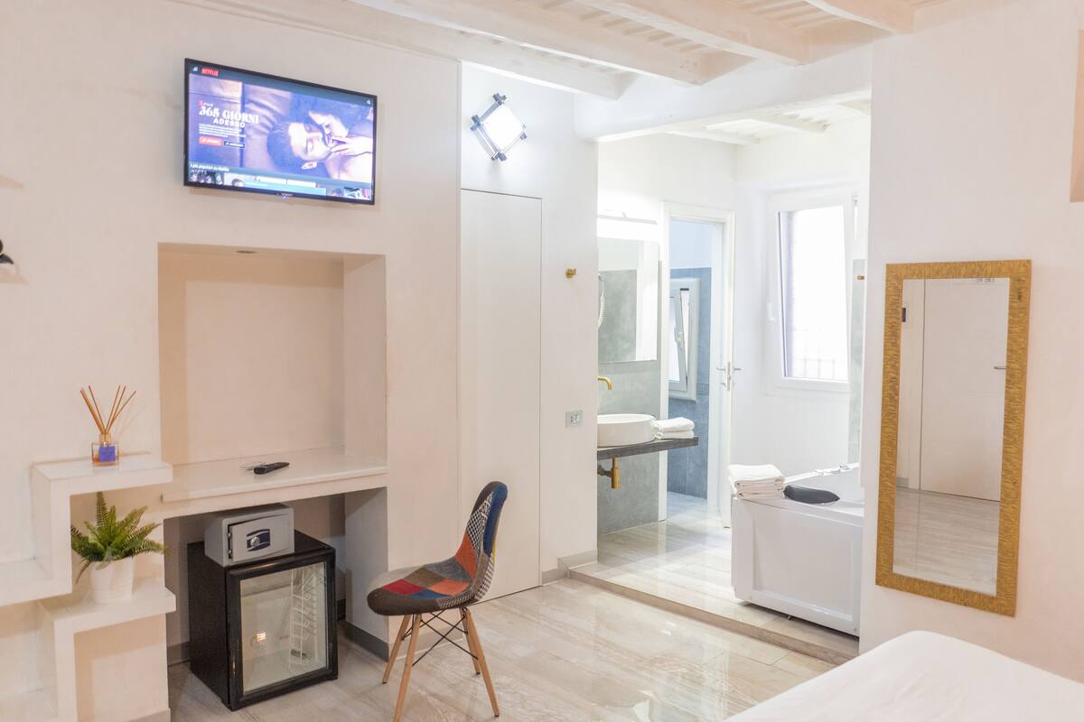 A modern hotel room with a TV mounted on the wall, a small desk with a computer, a chair, a bed, and a bathroom with a mirror and sink.
