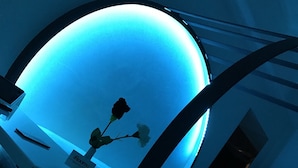 Una habitación con un llamativo elemento circular iluminado en azul, una cama con ropa blanca, una mesita con un florero y un perchero.