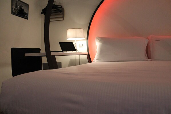Quarto de hotel com cama, escrivaninha, laptop, cadeira e luminária.