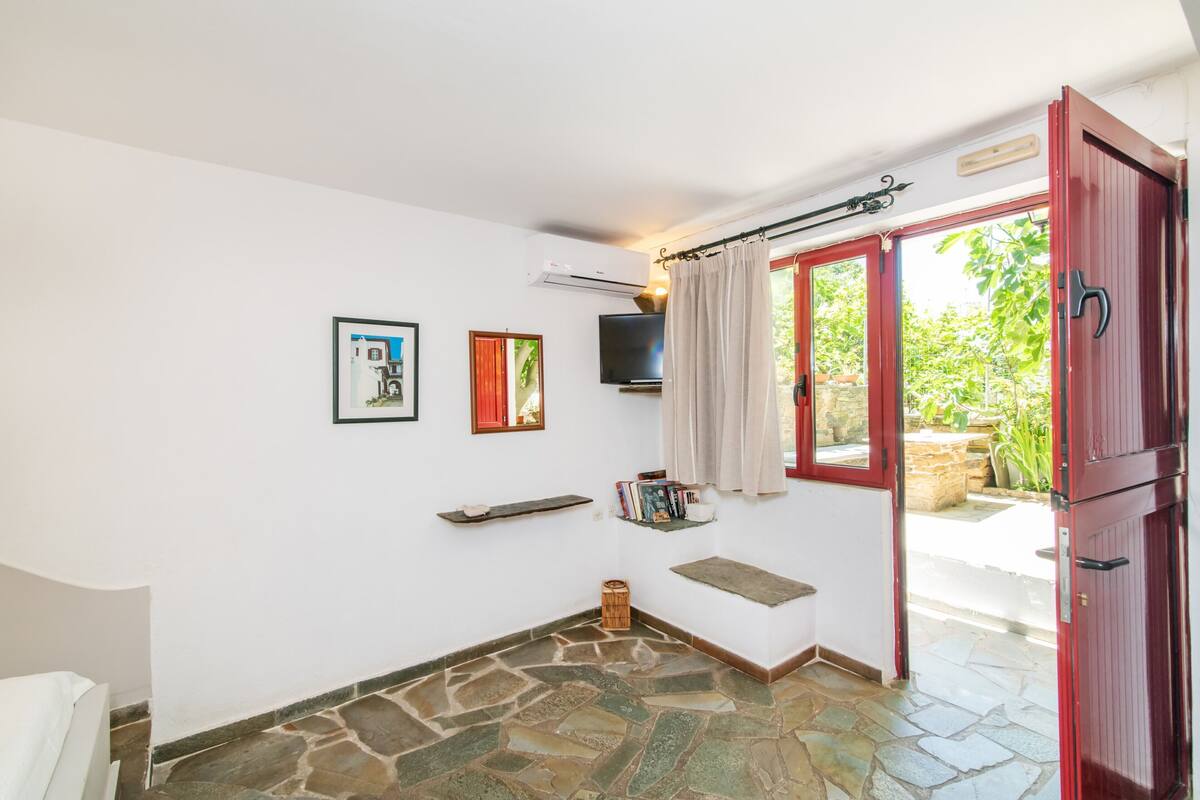 A room with a stone floor, a bed, a TV, a red door, and a view of greenery outside.