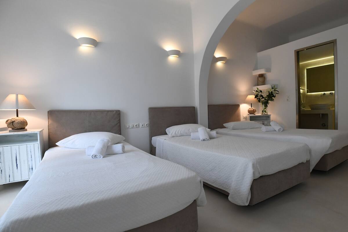 A hotel room with two beds, a nightstand with a lamp, a vase with flowers, and a mirror with robes hanging.