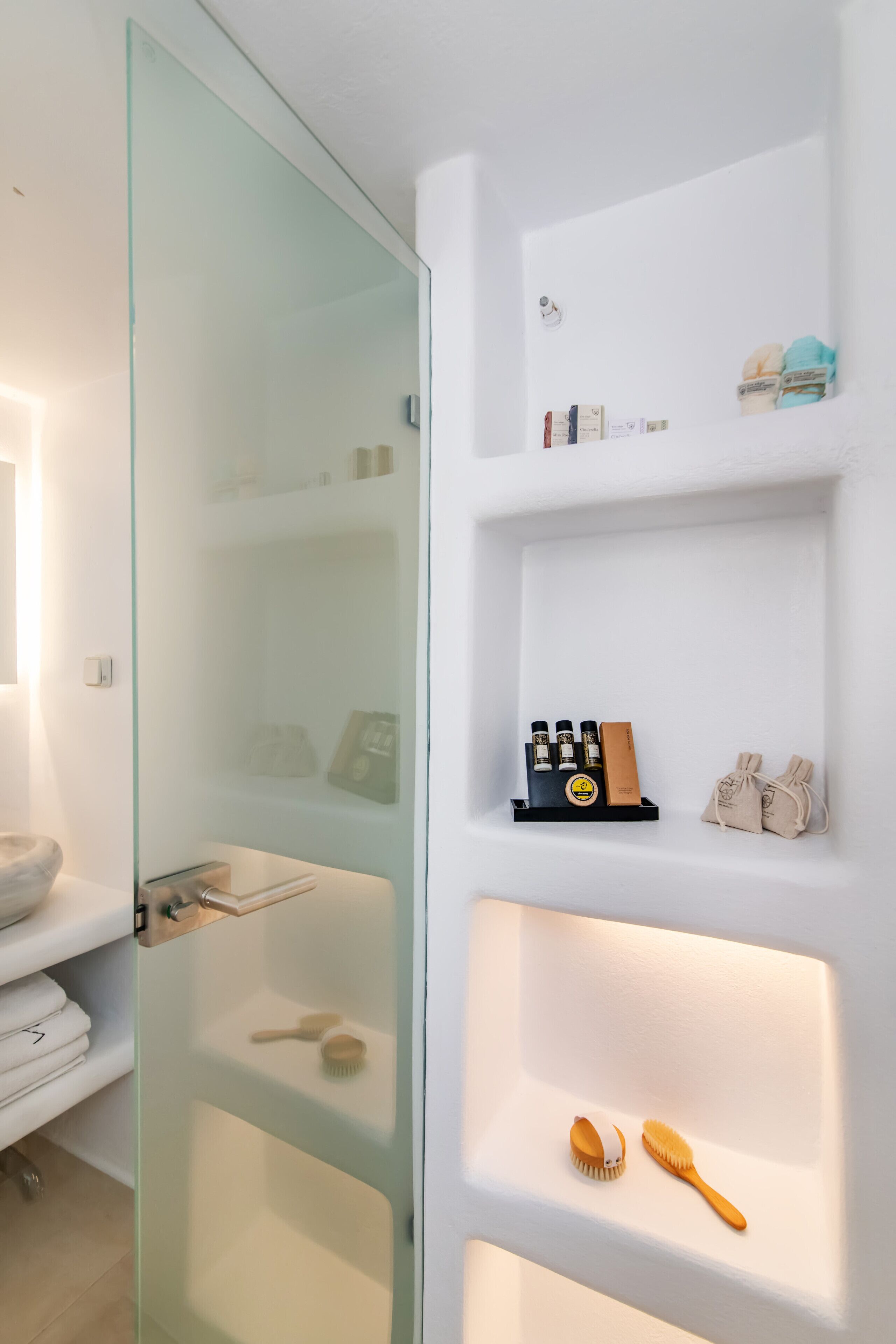 A bathroom with a glass shower door, a white sink, and shelves with toiletries and a towel rack.