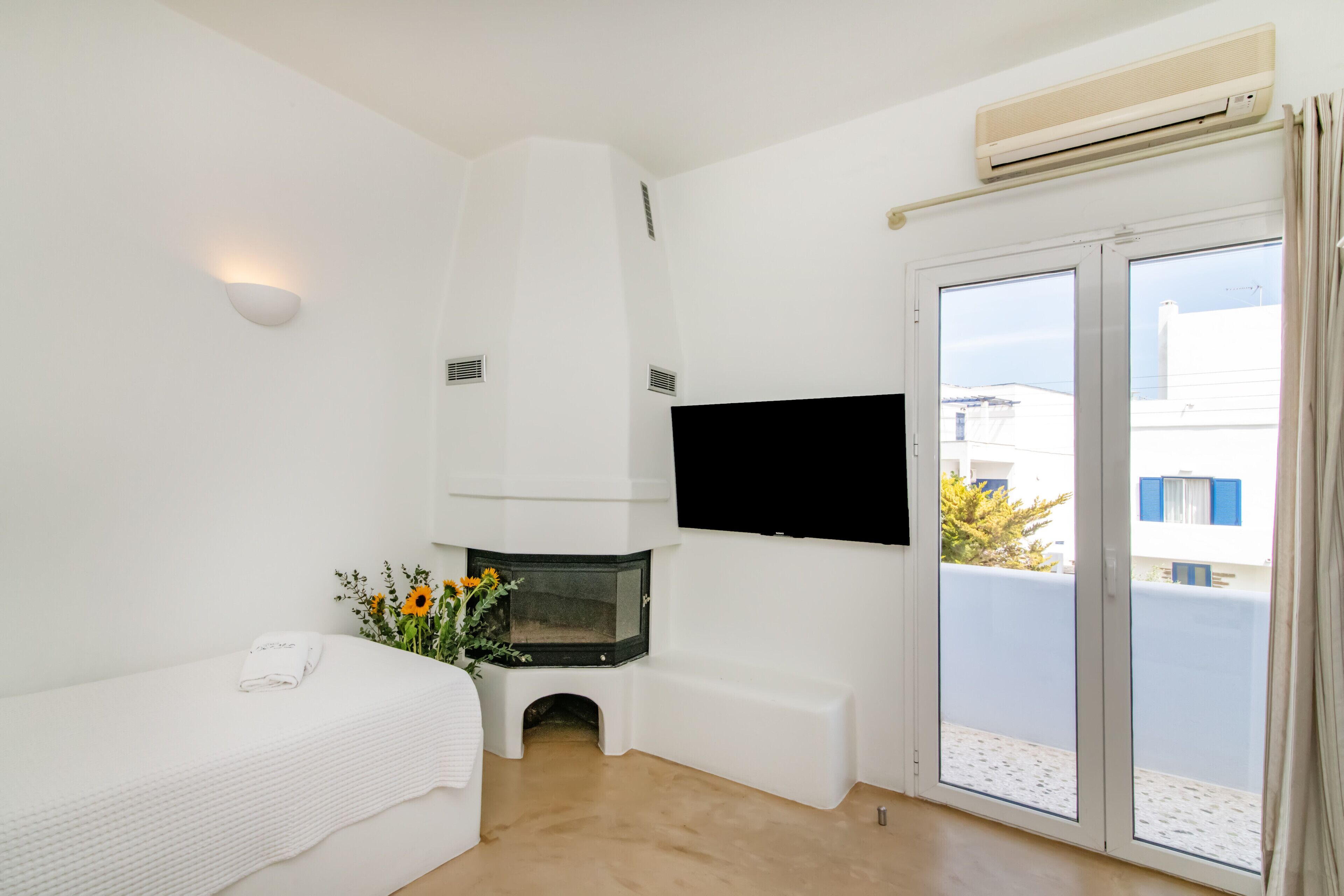 A modern hotel room with a flat-screen TV mounted on the wall, a built-in fireplace, a balcony with a view of the city, and a neatly made bed with a towel.