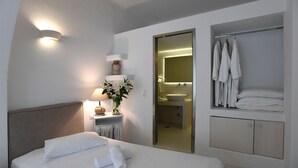 Habitación de hotel con cama, lámpara de noche, un florero con flores, baño con lavabo y espejo, y un perchero para toallas.