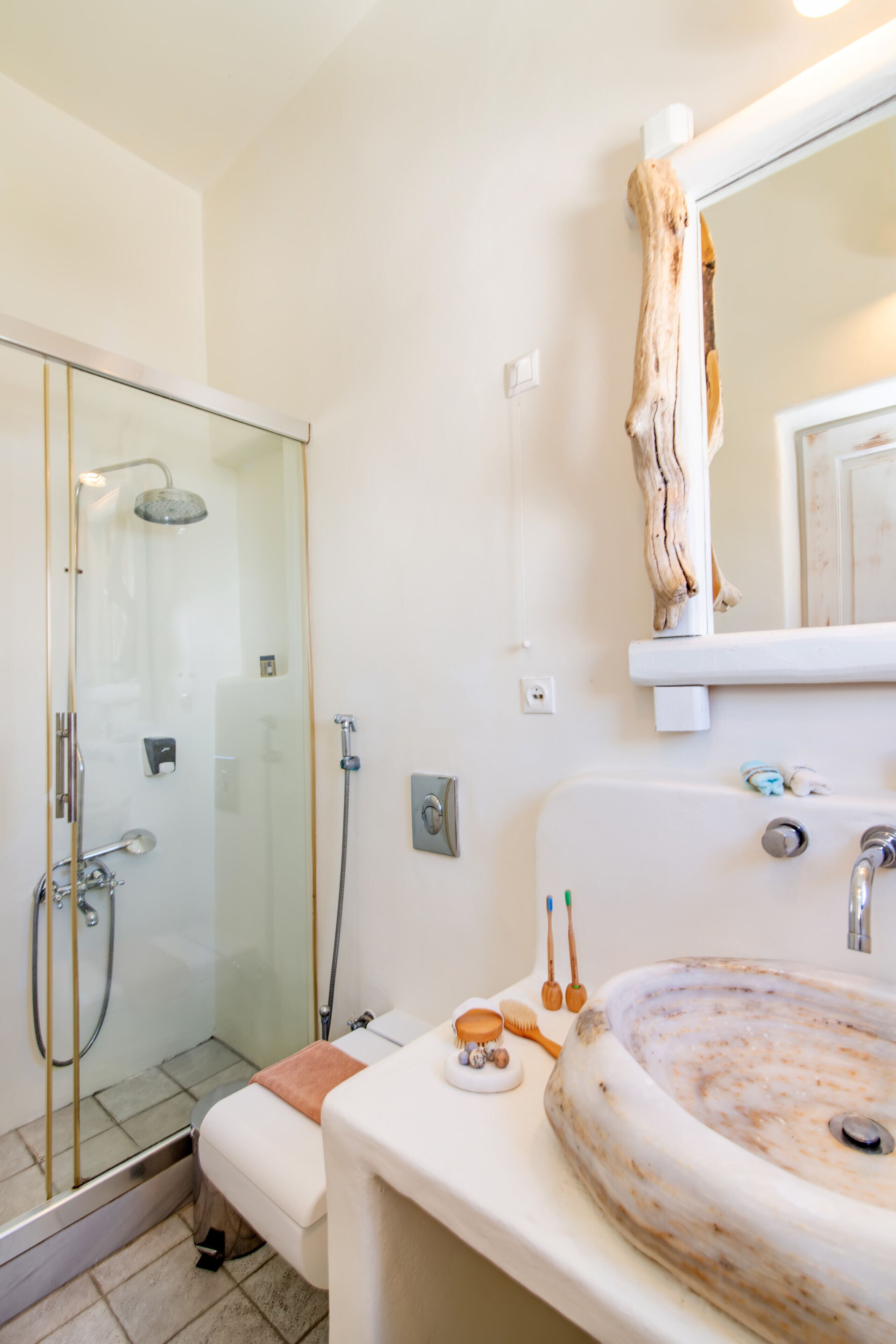A bathroom with a glass-enclosed shower, a white sink with a marble basin, and a wooden shelf above the mirror.
