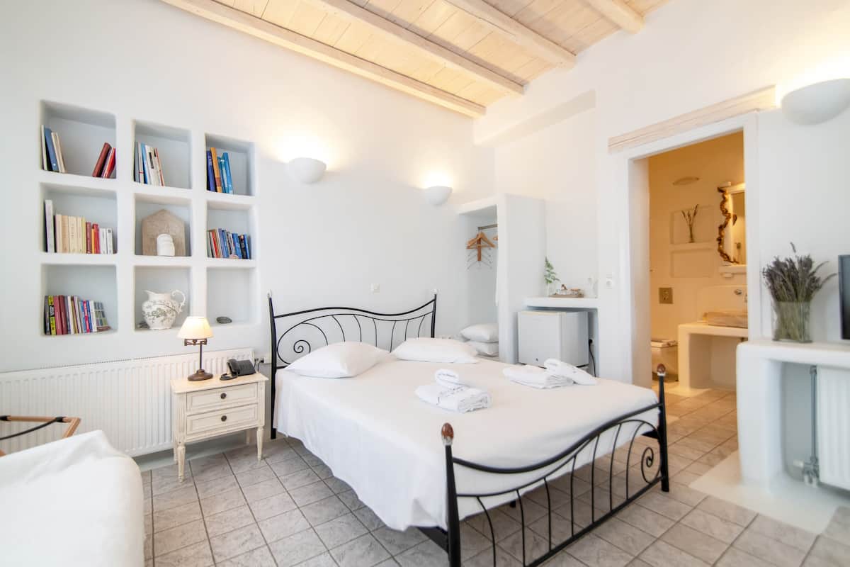 A neatly arranged bedroom with a bed, bedside tables, a bookshelf, and a bathroom visible through an open door.