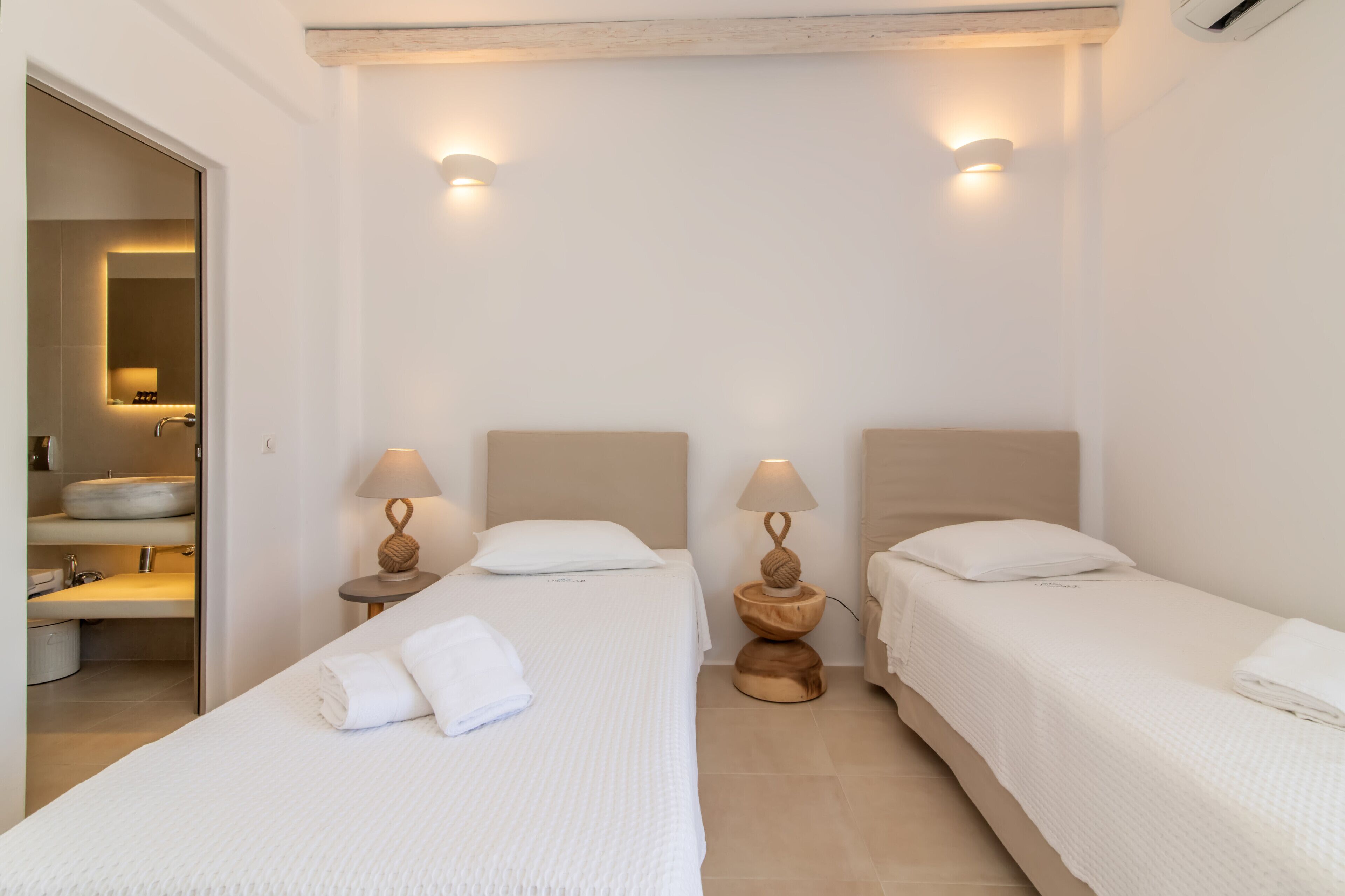 Habitación de hotel con dos camas individuales, baño con lavamanos y espejo, y un pequeño espacio de descanso con mesa y sillas.