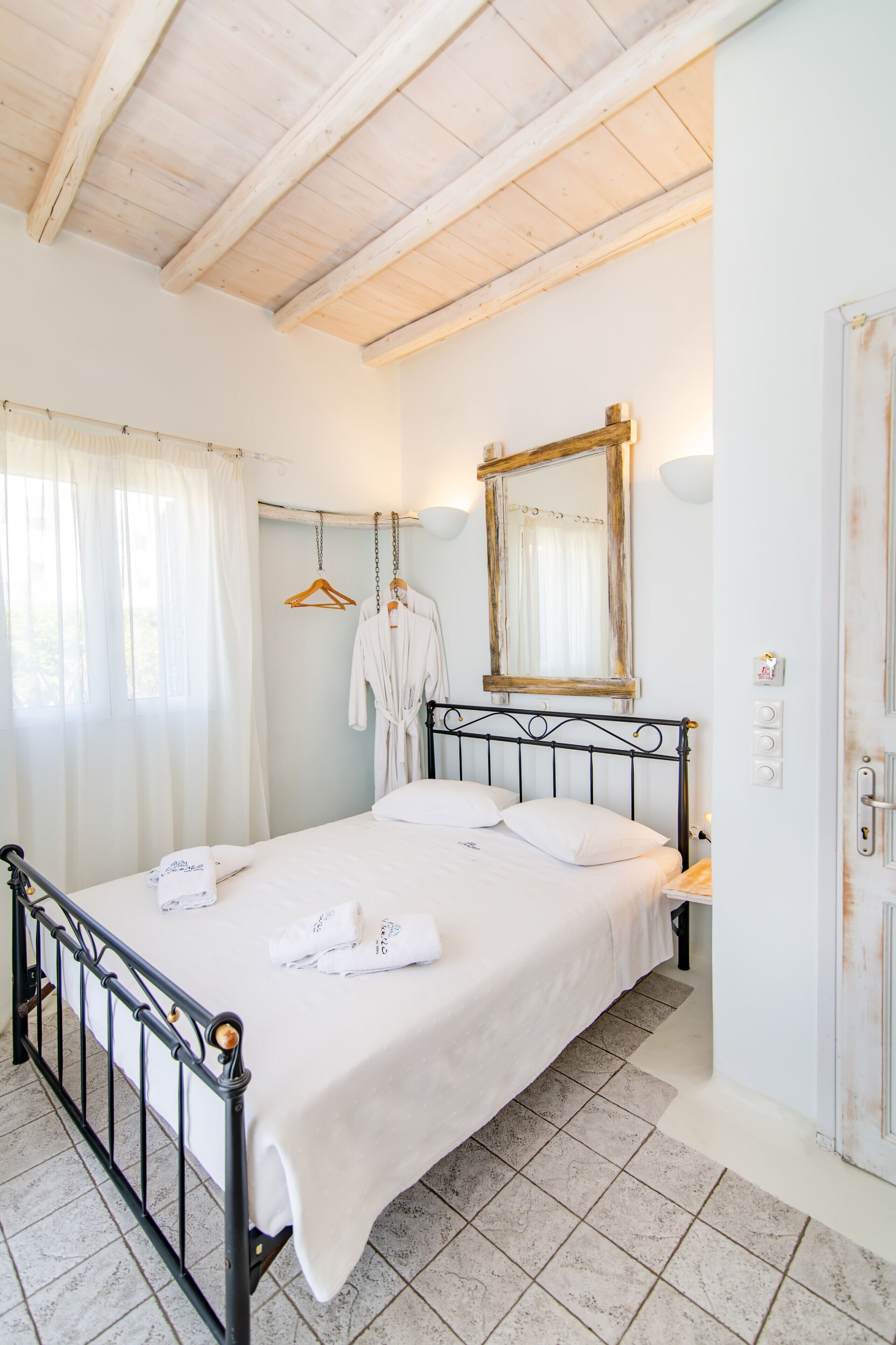A bedroom with a bed, white curtains, a wooden ceiling, a mirror, and a door.
