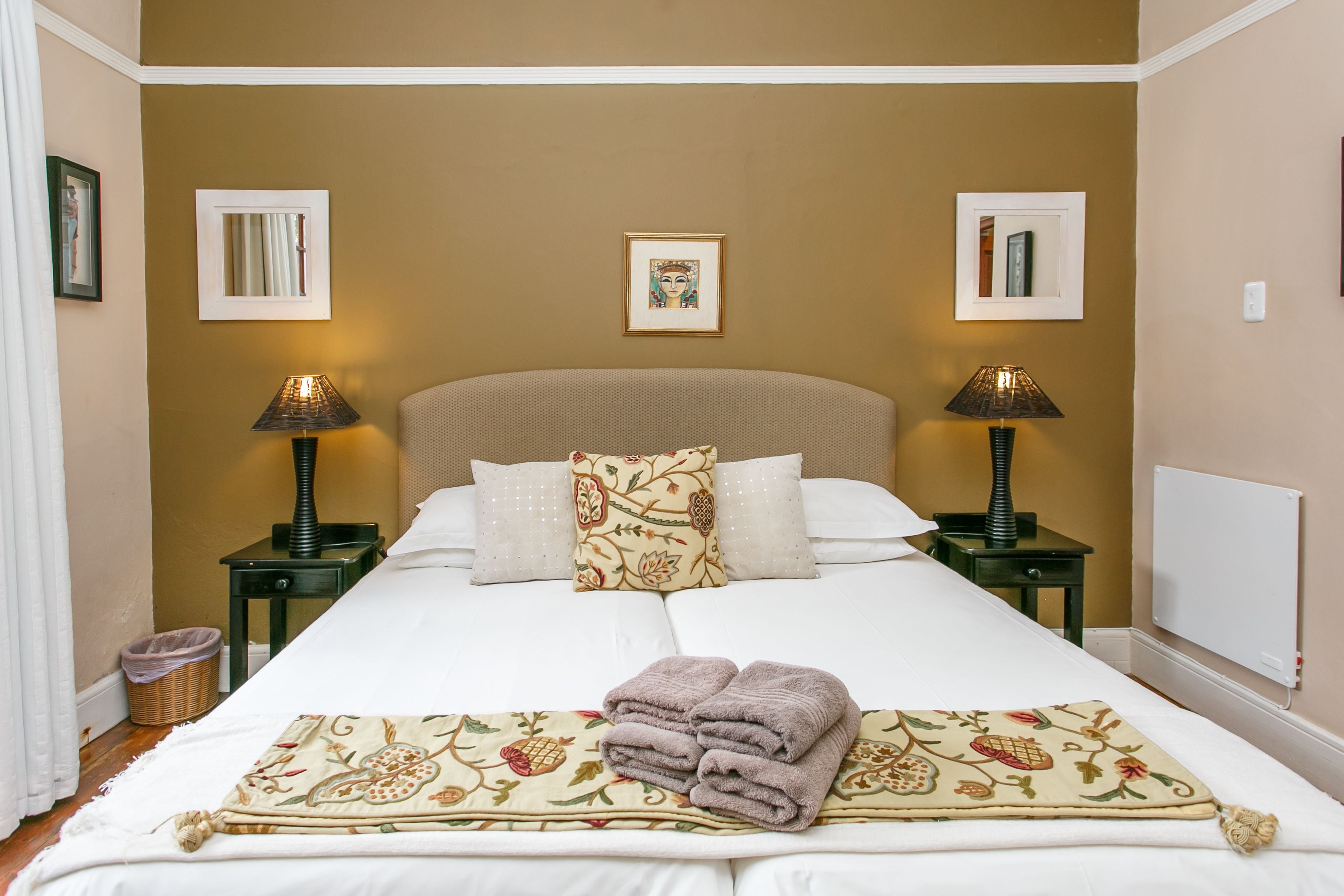 A neatly made bed with a floral throw blanket and towels, two bedside lamps, and framed artwork on the walls.