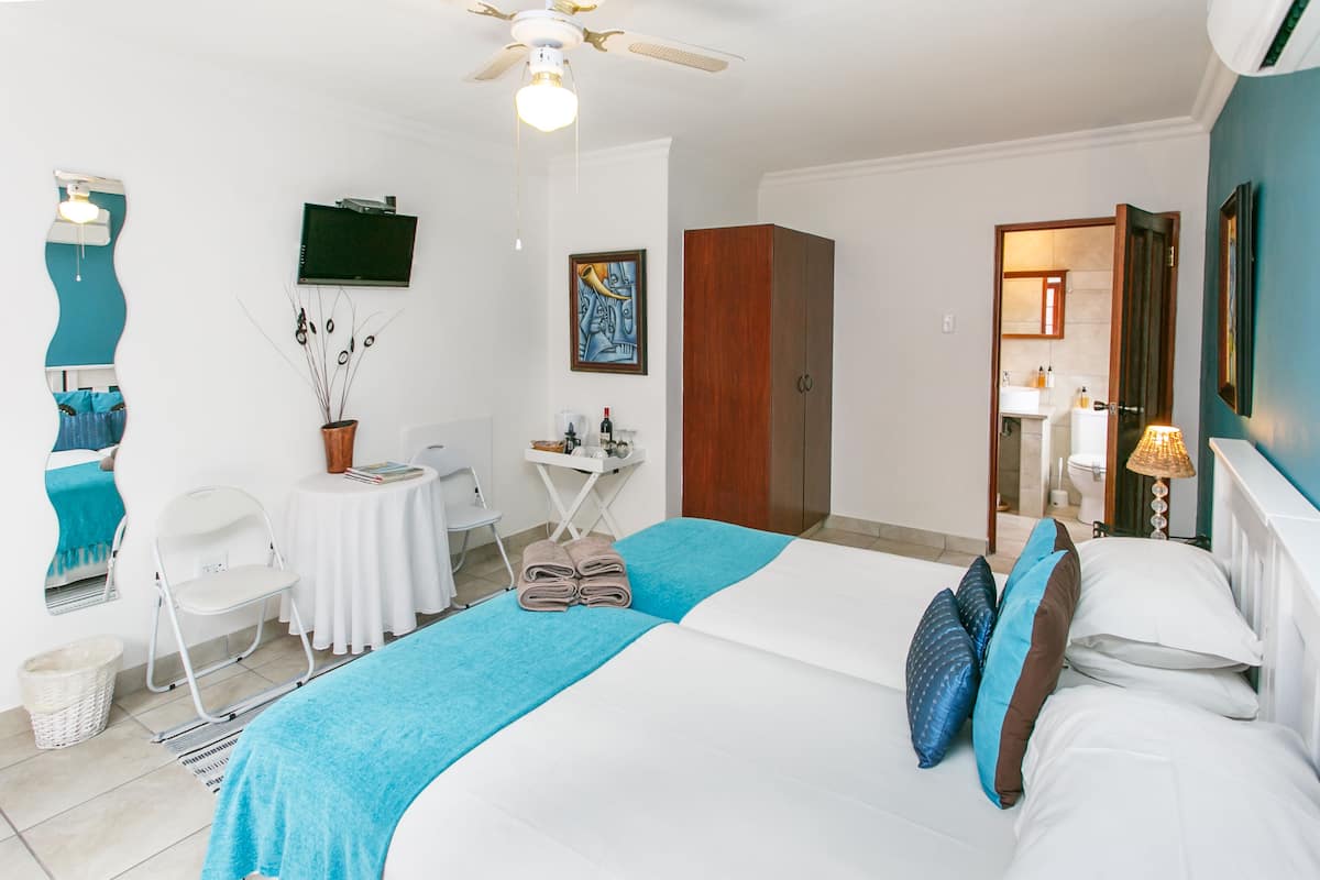 Ein Hotelzimmer mit Bett, Deckenventilator, kleinem Tisch mit Stuhl, Zimmerpflanze und angrenzendem Badezimmer.