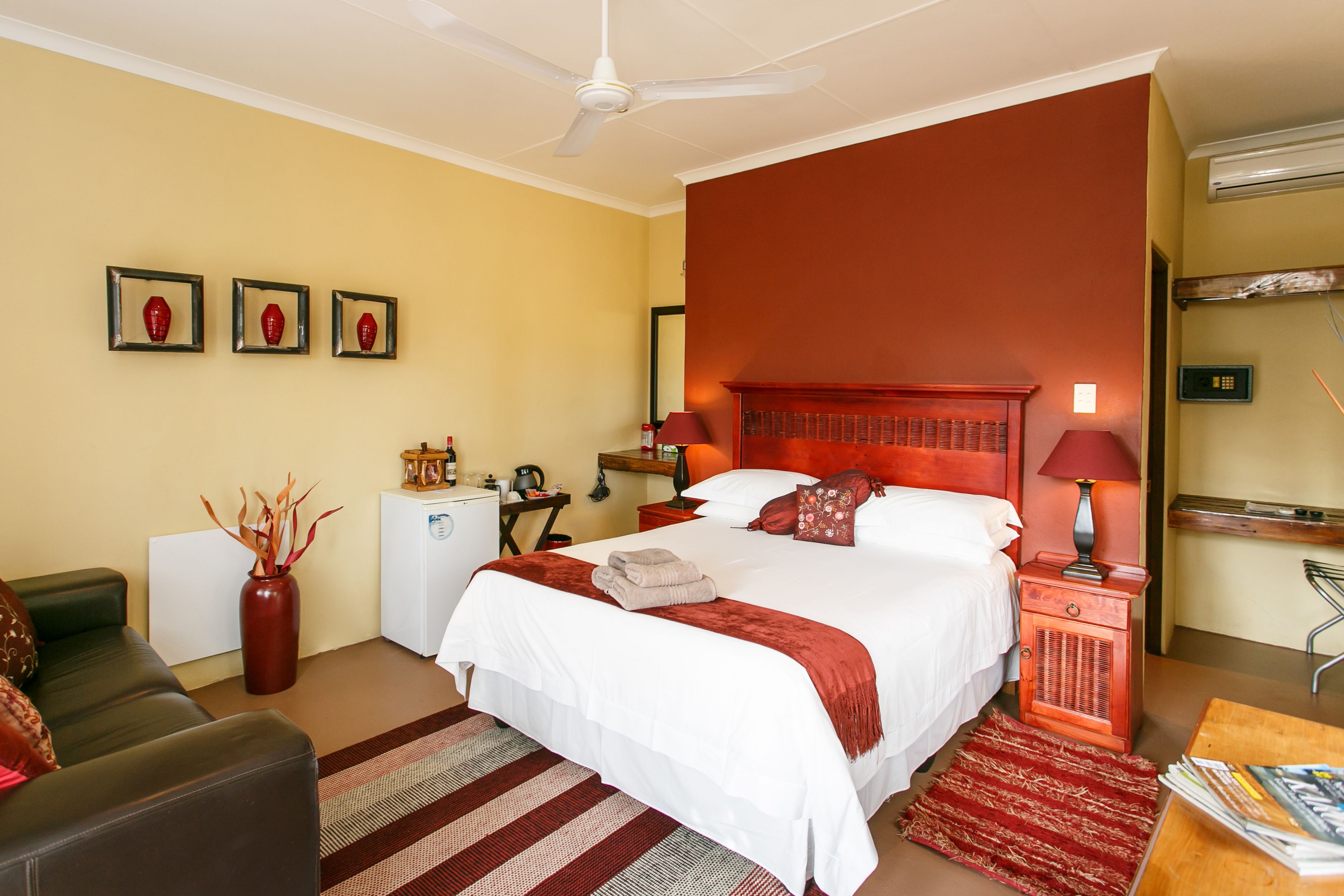 A hotel room with a large bed, a desk, a chair, a small table, a red vase, and a ceiling fan.
