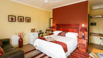 A hotel room with a large bed, a desk, a chair, a small table, a red vase, and a ceiling fan.