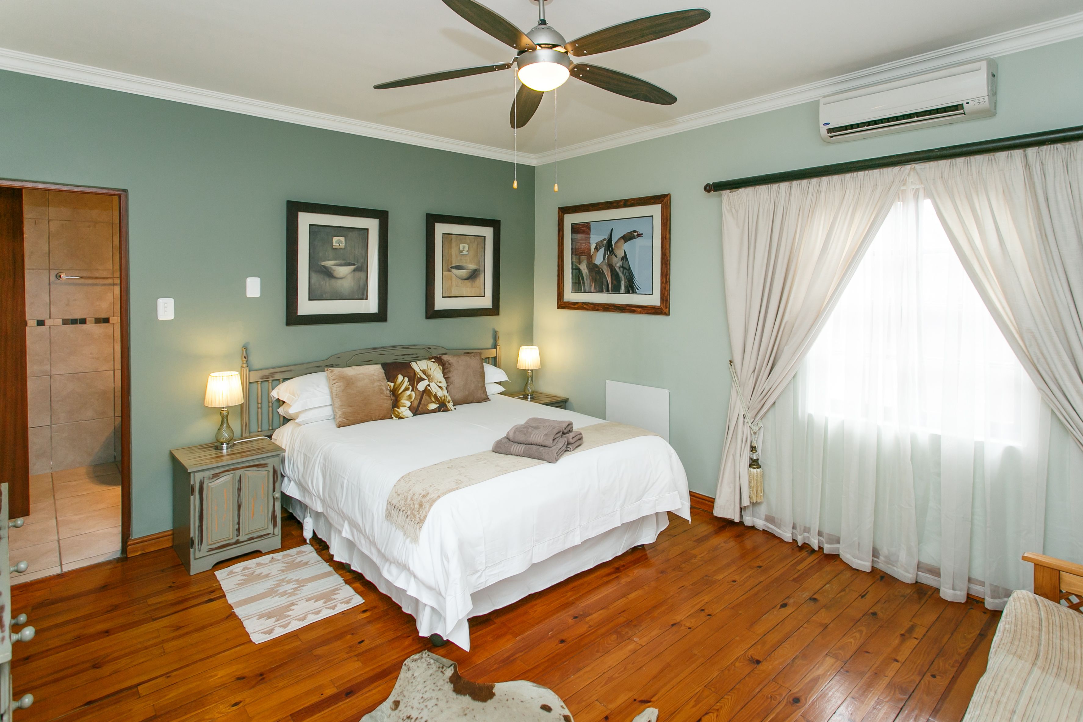 A bedroom with a large bed, wooden flooring, a ceiling fan, and framed artwork on the walls.