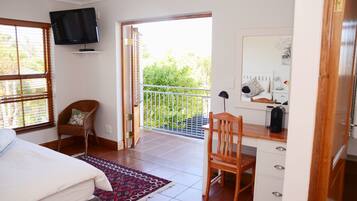 A bedroom with a bed, a desk, a chair, a television, and a balcony with a view of greenery.