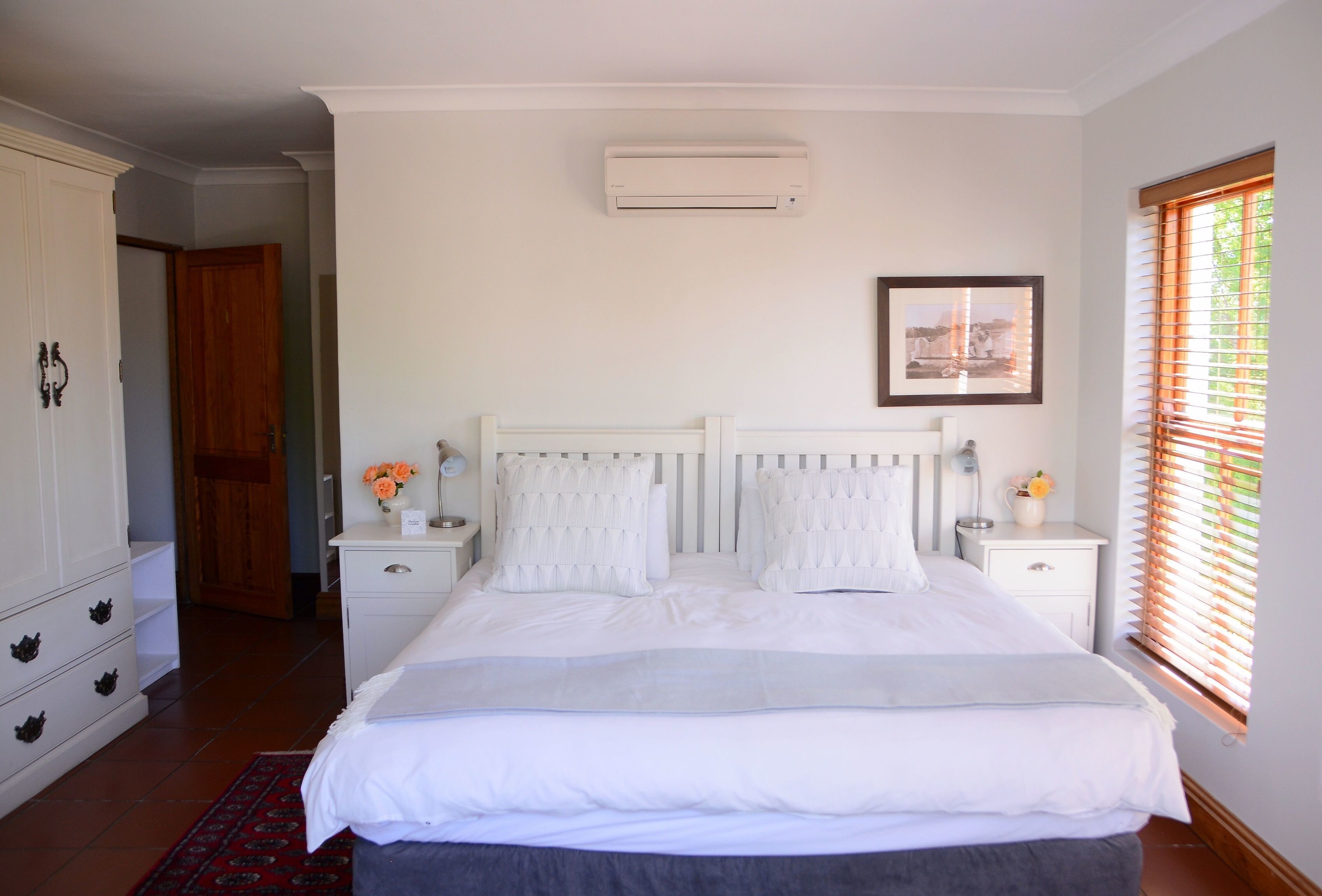 Une chambre à coucher avec un lit, des tables de chevet blanches, une table de chevet, un climatiseur fixé au mur et une fenêtre avec des stores.