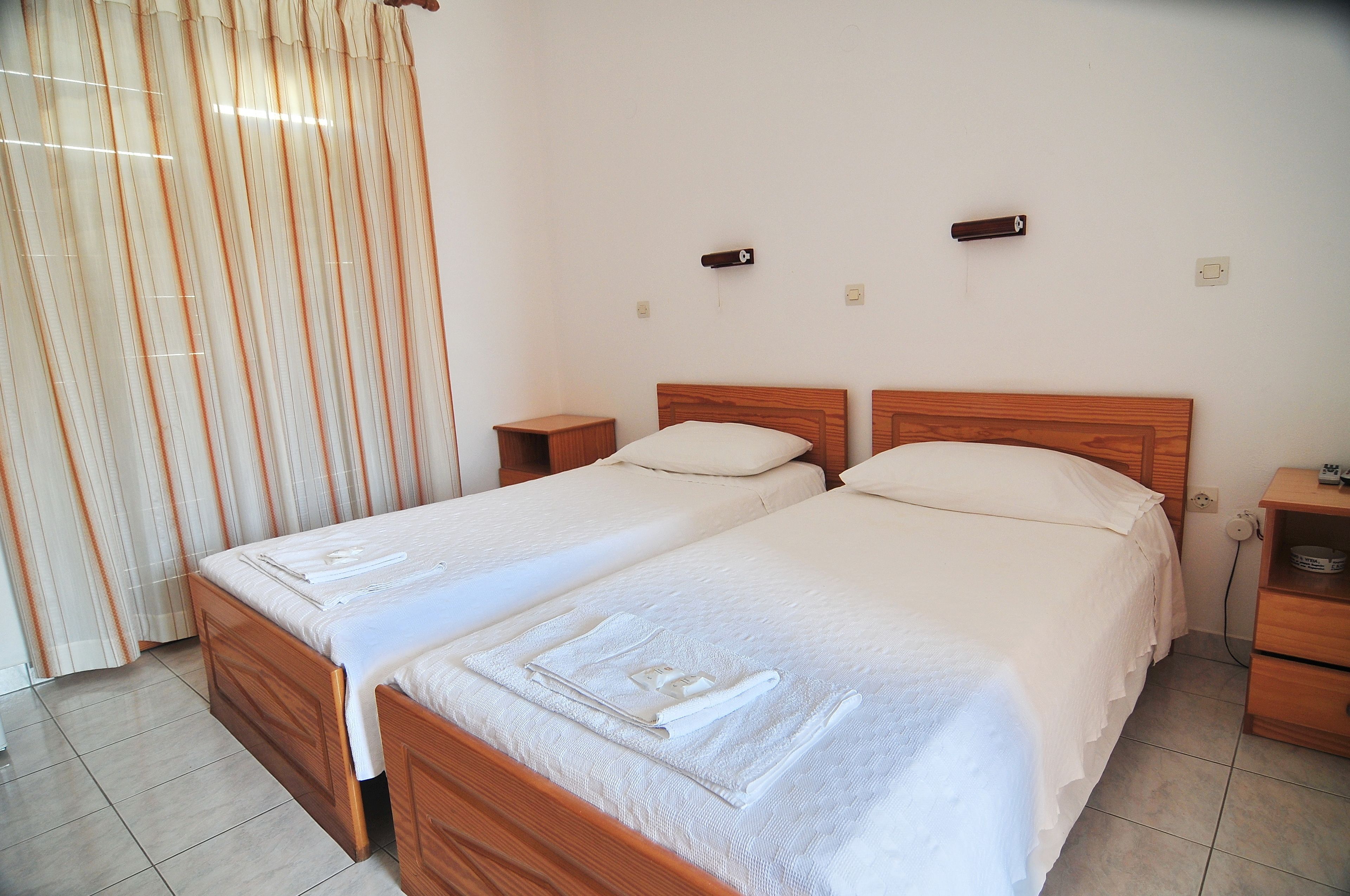 A double bed with white linens and a wooden headboard, a nightstand with a lamp, and a window with curtains.