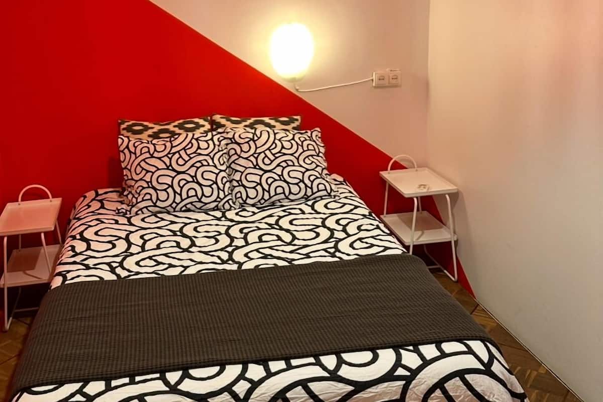 A bed with a patterned bedspread and pillows, a bedside table, a nightstand, and a wall-mounted light.