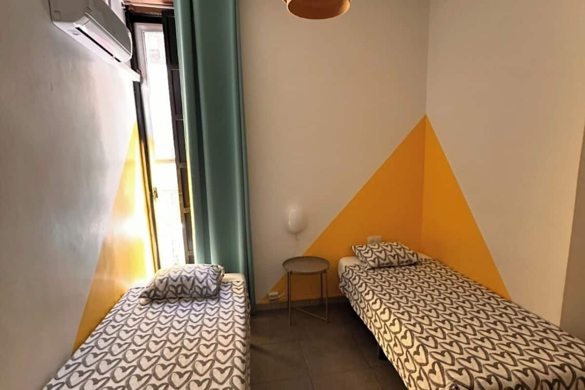 A small, brightly lit room with two single beds, a small table, and a hanging light fixture.